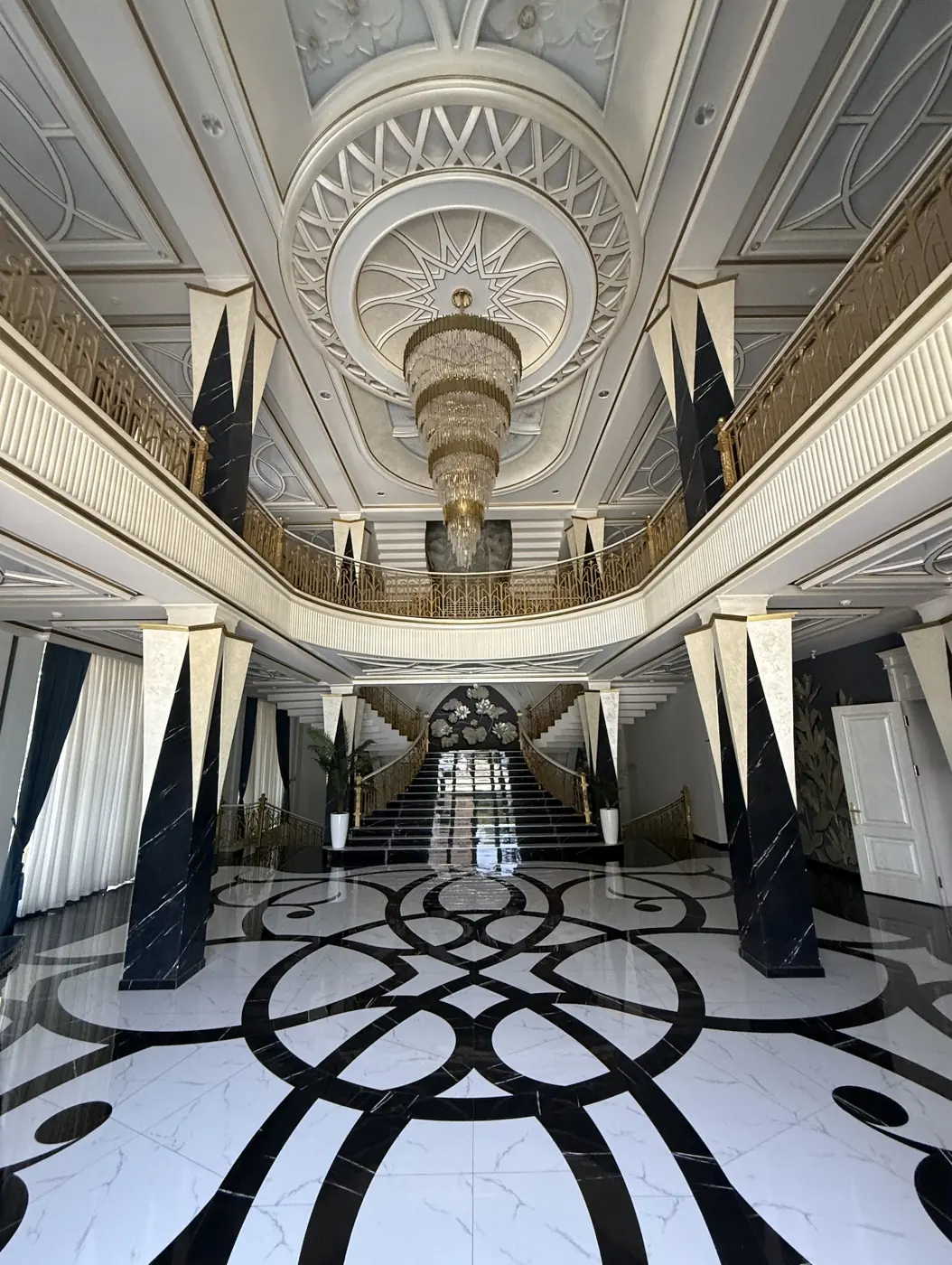 The grand interior of Saroy Event Hall with a crystal chandelier, sweeping staircase, gold railings, and patterned marble floor